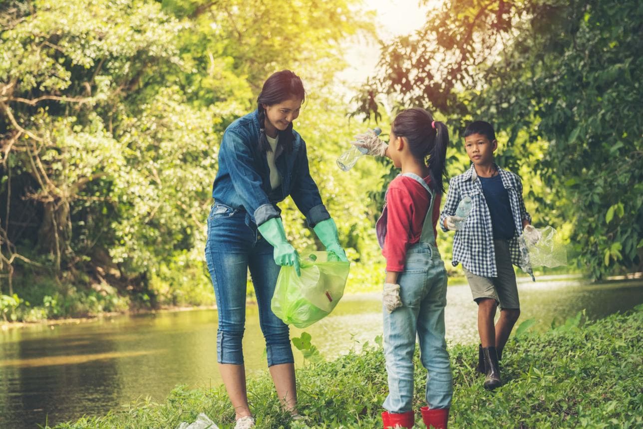 O voluntară și doi copii adună sticle de plastic pe malul unui pârâu, în lumina caldă a după-amiezii.